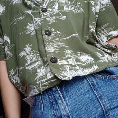 Cropped short-sleeve shirt in lightweight khaki green fabric with white tropical print. Revere collar, black button closure at front, loose fit. Soft fabric, comfortable for summer. In good overall condition with no visible defects.