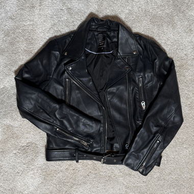 Veste en cuir courte noire avec fermeture éclair à l'avant, plusieurs poches zippées, zips aux manches et ceinture à la taille. Doublure intérieure noire.