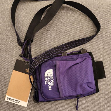 Pochette porte-bouteille The North Face violette avec bandouliere reglable, poche zippee et empiècements en maille. Utilisable en bandouliere ou en accessoire de marche/randonnee. Neuf
